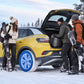 A bright yellow compact SUV with mounted snow socks is parked in the snow with three people around it, one loading gear into the open trunk and two holding snowboards. 
